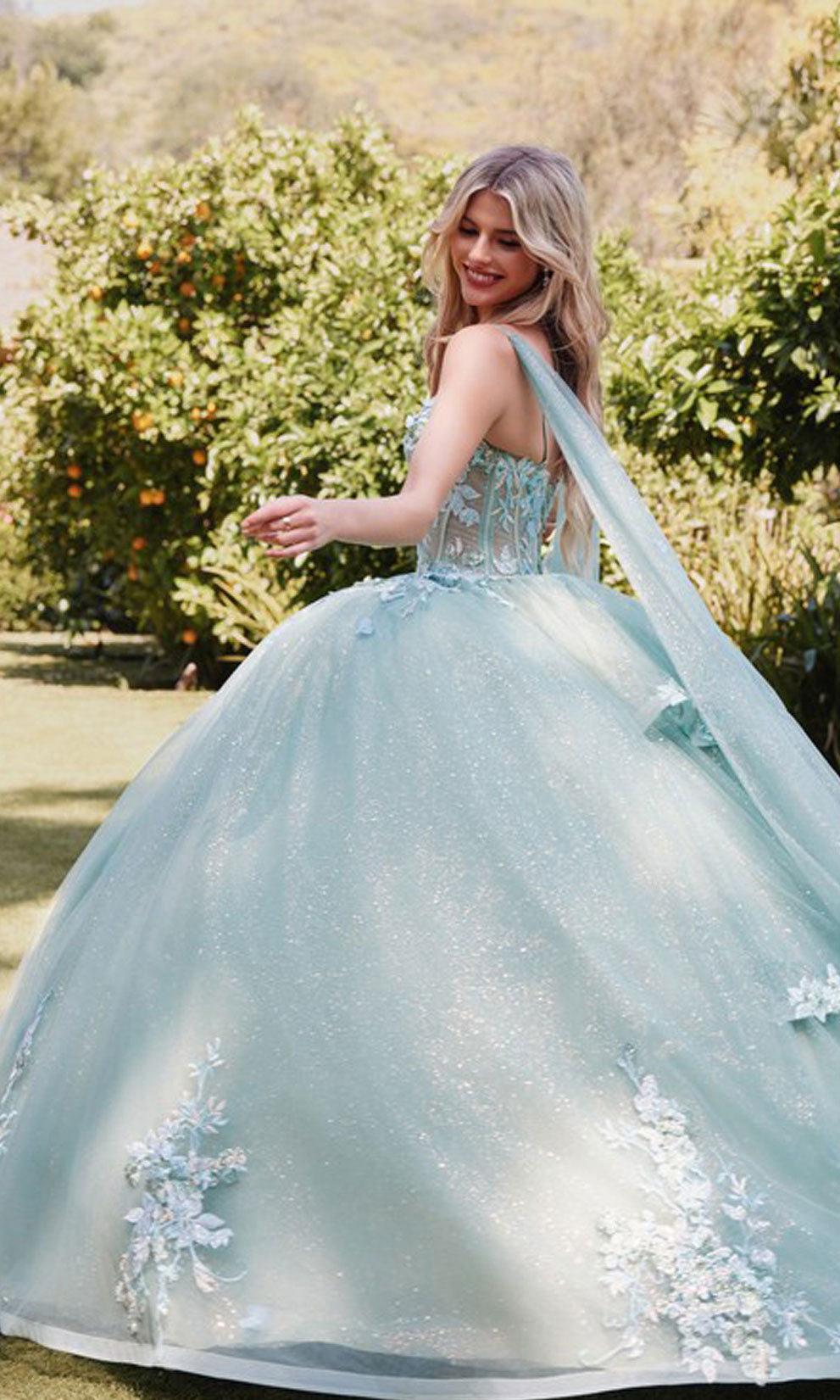 Outdoors in a garden, a person turns and smiles, wearing the Sage Green Quinceanera Dress: Juliet JT1466H—a gown with a sheer corset bodice and glittering skirt, its elegant details catching the sunlight.